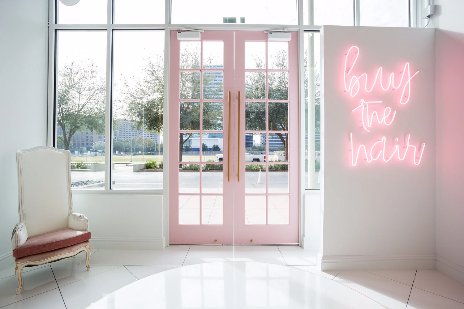 Salon entrance with large pink double doors, a white high-back chair, and a pink neon sign reading “buy the hair.”